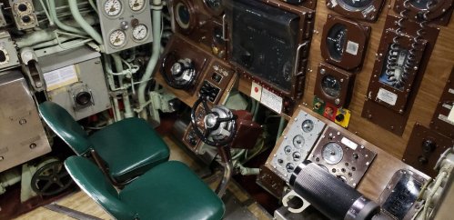 Control panel of a submarine featuring gauges, levers, and a steering wheel, with green chairs in the foreground.