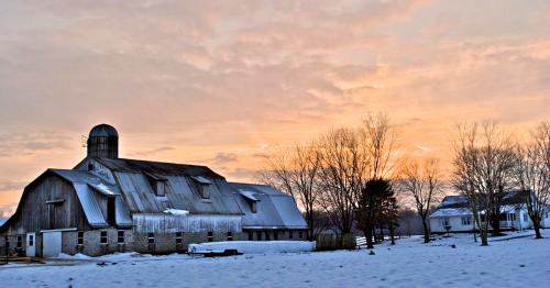 A snowy farm scene at sunset, featuring a weathered barn and trees against a colorful sky.