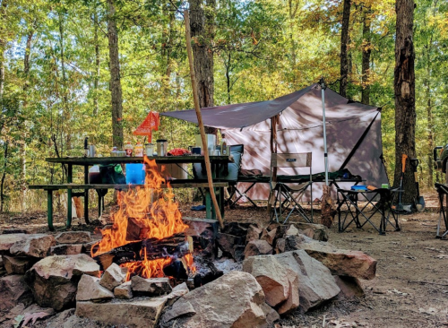 A cozy campsite with a fire pit, flames, a tent, and picnic table surrounded by trees in a colorful autumn setting.