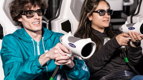 Two people wearing 3D glasses hold gaming guns while seated in a futuristic ride or game setup.