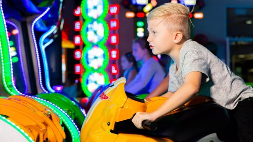 A young boy rides a yellow arcade motorcycle, focused and enjoying the colorful lights of the game room.