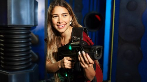 A young woman smiles while holding a futuristic laser gun in a colorful gaming environment.