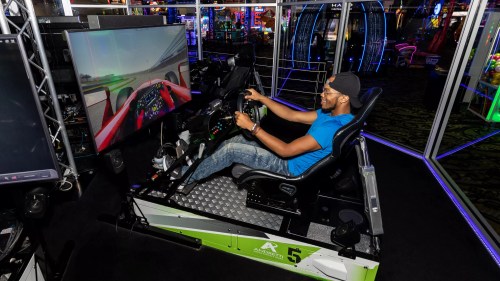 A person sits in a racing simulator, focused on the screen displaying a racing game in a vibrant arcade setting.