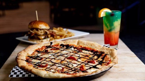 A delicious pizza with toppings and balsamic drizzle, alongside a burger, fries, and a colorful drink.