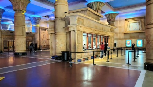 Interior of a grand, Egyptian-themed space with columns, decorative walls, and a ticket counter. People are in line.