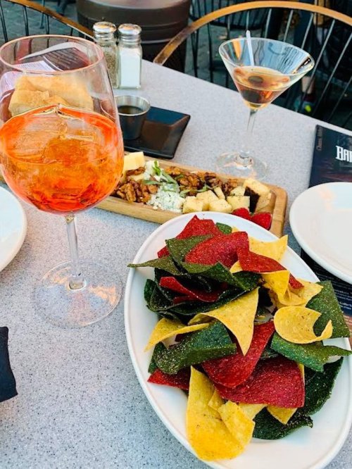 A plate of colorful tortilla chips in red, green, and yellow, with a drink and a cheese platter in the background.