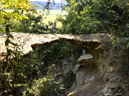 A natural stone arch surrounded by greenery, overlooking a scenic valley below.