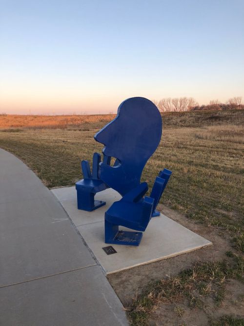 A large blue sculpture of a head and two chairs, set against a grassy landscape at sunset.