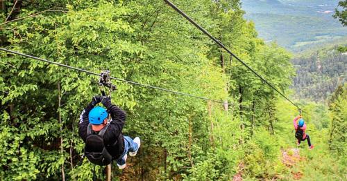 Two people ziplining through a lush green forest, with a scenic view of mountains in the background.