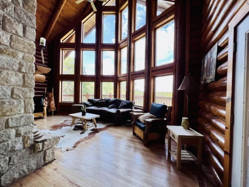 Cozy log cabin interior with large windows, wooden floors, and a stone fireplace, featuring a sofa and coffee table.