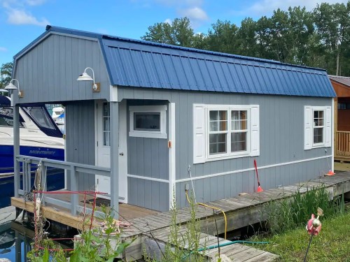 A small gray houseboat with a blue metal roof, white shutters, and a wooden deck, docked by a marina.