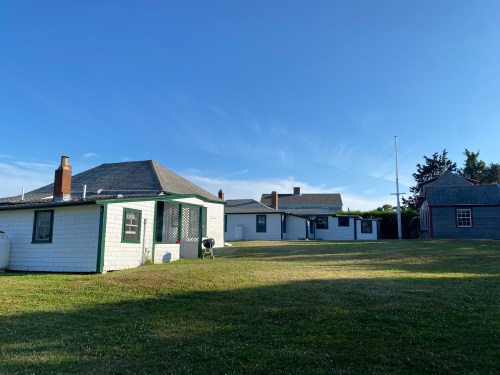 A sunny outdoor view of several small, white houses with green trim on a grassy lawn under a clear blue sky.