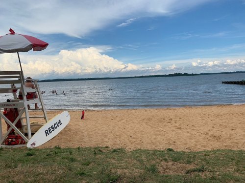 A sandy beach with a lifeguard stand, red and white umbrella, and people swimming in the water under a cloudy sky.