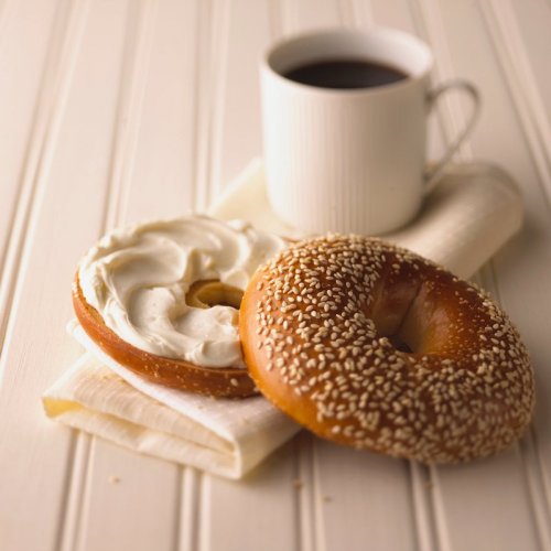 A sesame bagel and a cream cheese-filled bagel beside a cup of coffee on a white table.