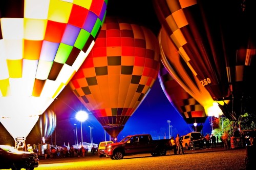Colorful hot air balloons illuminated at night, with trucks and spectators in the foreground.