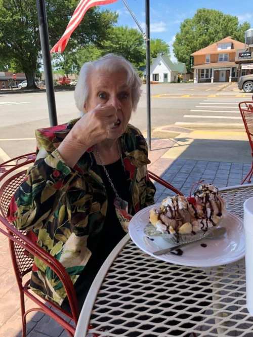An elderly woman enjoys a large banana split dessert while sitting at an outdoor café table.