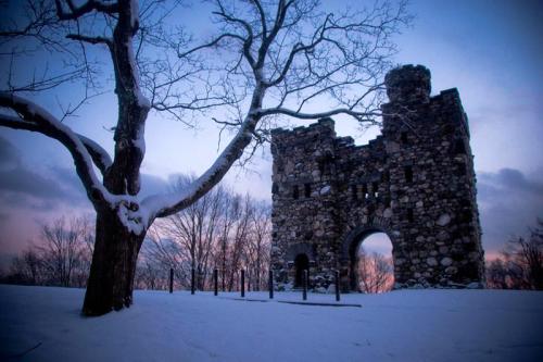 A snow-covered stone ruin stands against a twilight sky, with a bare tree nearby and soft clouds in the background.