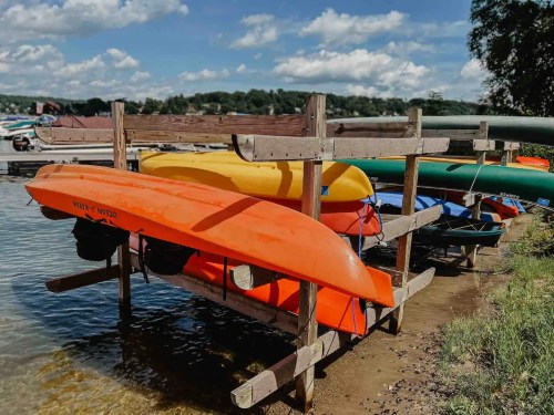 Colorful kayaks are stacked on a wooden rack by the water, with a scenic view of the lake and blue sky in the background.