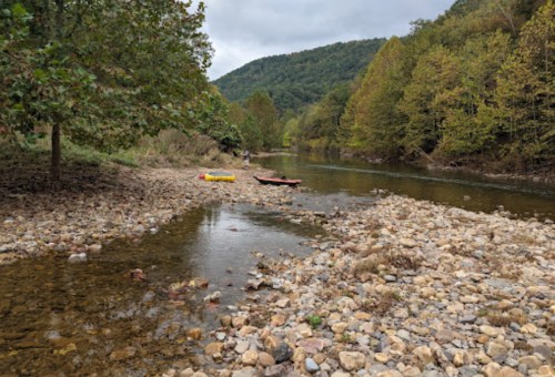 A serene river scene with rocky banks, trees lining the shore, and a gentle flow of water under a cloudy sky.