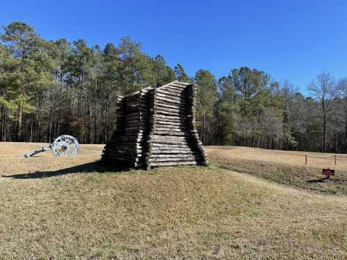 A wooden structure made of logs stands in a grassy field, with trees in the background and a cannon nearby.