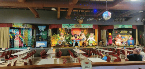 A colorful stage with animated characters and decorations, set in a dining area with empty tables and chairs.