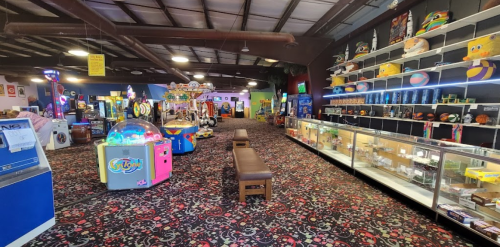 A colorful arcade interior featuring games, prizes, and a carpeted floor, with benches and display cases along the walls.