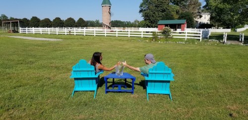 A couple sits in blue chairs, toasting with drinks at a table in a grassy field with a windmill in the background.