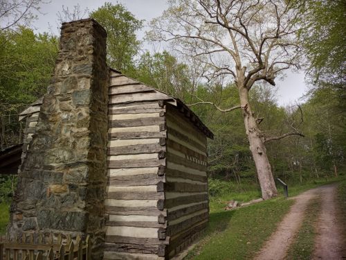 A rustic log cabin with a stone chimney, surrounded by trees and a dirt path leading into the woods.