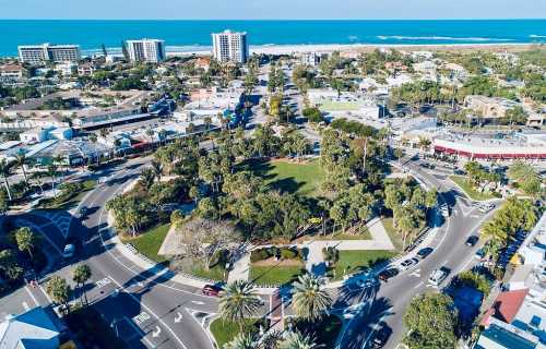 Aerial view of a coastal town with a roundabout, palm trees, and buildings near the beach and ocean.