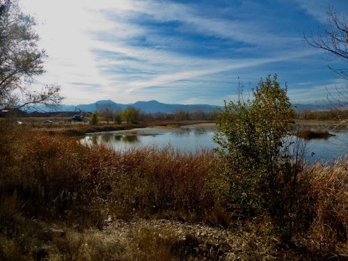 A serene landscape featuring a calm lake, autumn foliage, and distant mountains under a blue sky with wispy clouds.