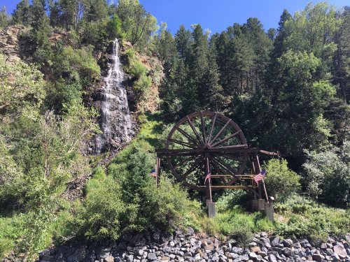 A wooden water wheel beside a waterfall, surrounded by lush greenery and trees.