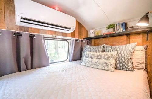 Cozy interior of a camper with a bed, air conditioning unit, and shelves filled with books and decor.