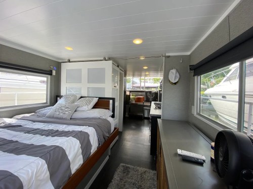 Cozy interior of a modern houseboat featuring a bed, kitchenette, and large windows with views of the marina.