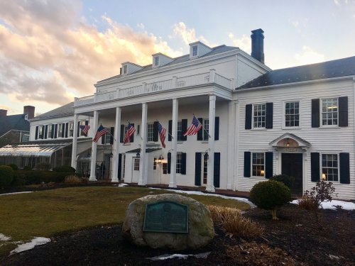 Historic white building with columns, flags, and a plaque, surrounded by landscaped grounds and a sunset sky.