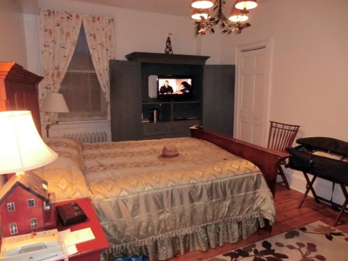 Cozy bedroom with a bed, TV on a cabinet, a chair, and a lamp, featuring floral curtains and wooden flooring.