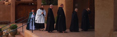 A group of monks in black robes walking towards a doorway in a serene outdoor setting.