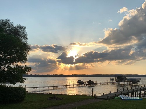 Sunset over a lake with rays of light breaking through clouds, boats docked along a wooden pier, and lush greenery nearby.