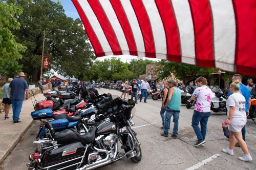 A bustling street filled with parked motorcycles and people walking, framed by an American flag in the foreground.