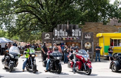 A group of motorcyclists gathers outside Buffalo Bill's Olde Fashion Soda Pop Co., with bikes parked in front.