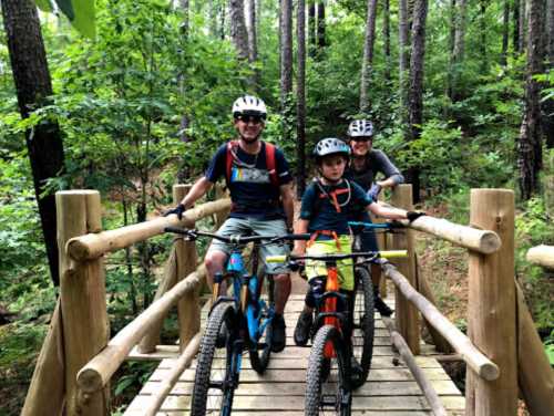 A family of three on mountain bikes stands on a wooden bridge in a lush, green forest.