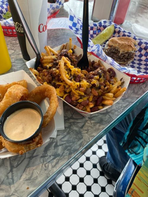 A plate of chili cheese fries topped with beans and cheese, alongside onion rings and a burger on a checkered table.