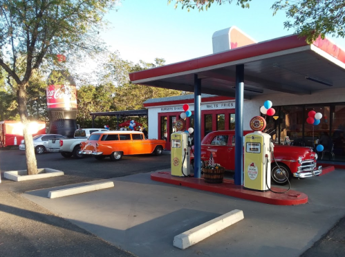 A retro diner with classic cars parked outside, featuring vintage gas pumps and festive decorations.