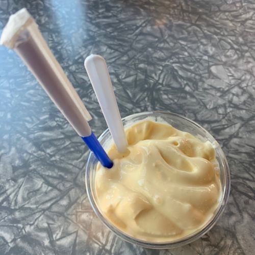 A cup of creamy yellow soft serve ice cream with two plastic spoons, resting on a textured gray surface.