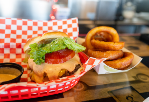 A juicy burger with lettuce, tomato, and sauce, served with a side of crispy onion rings in a red checkered basket.