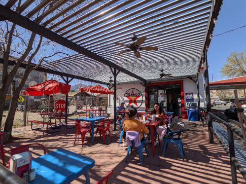 Outdoor dining area with colorful tables and umbrellas, people enjoying meals under a shaded pergola.