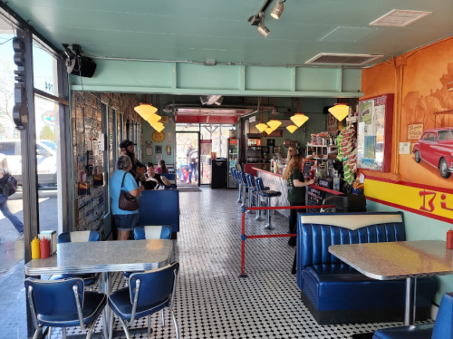 A retro diner interior with blue booths, a counter, and patrons. Bright lighting and colorful decor create a lively atmosphere.