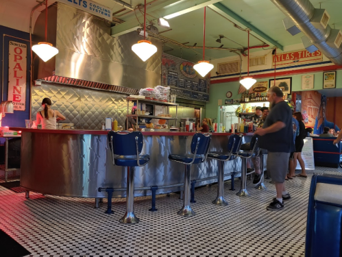 A retro diner interior with a chrome counter, blue stools, and patrons enjoying food in a vibrant, colorful setting.