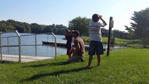A man and two children observe a lake from a grassy shore on a sunny day.
