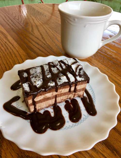A slice of layered chocolate dessert drizzled with sauce, served on a white plate next to a cup of coffee.