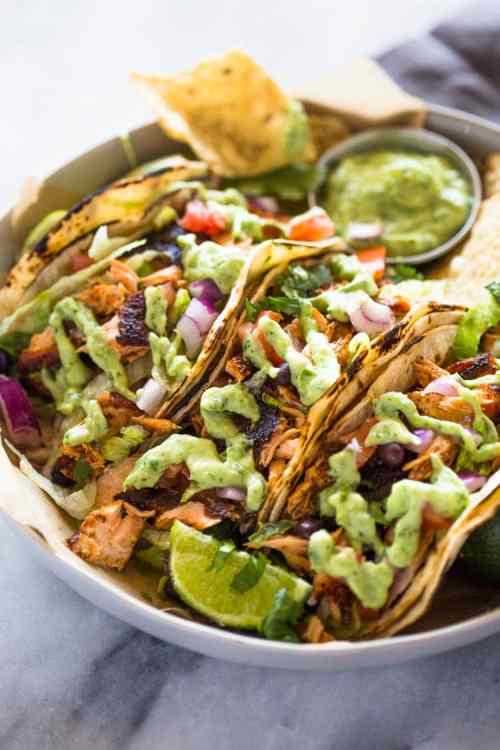 A plate of three grilled tacos topped with green sauce, onions, and tomatoes, served with chips and a side of salsa.
